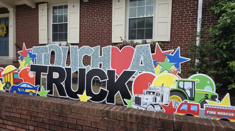 Colorful 'Touch a Truck' display with big glittery letters and toy vehicles in front of a brick house, stars and banners.